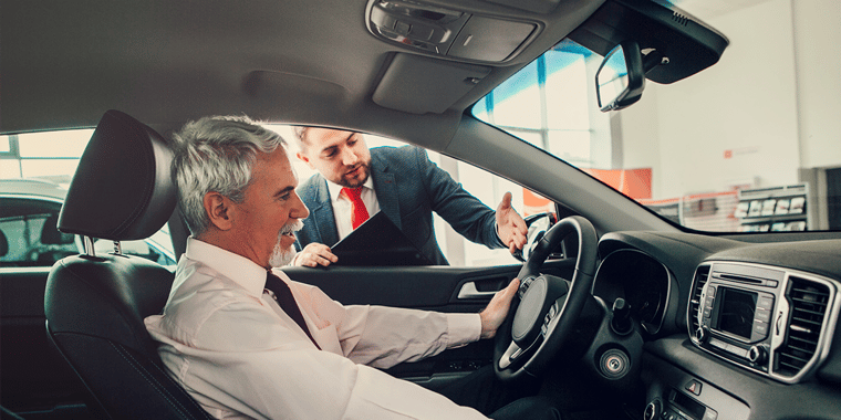 A car salesman talks to an older man, who's sitting in a car in a showroom. 