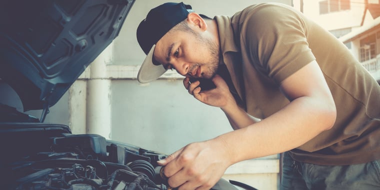 A man leans over and looks at something under the hood of his car, while calling someone on the phone.