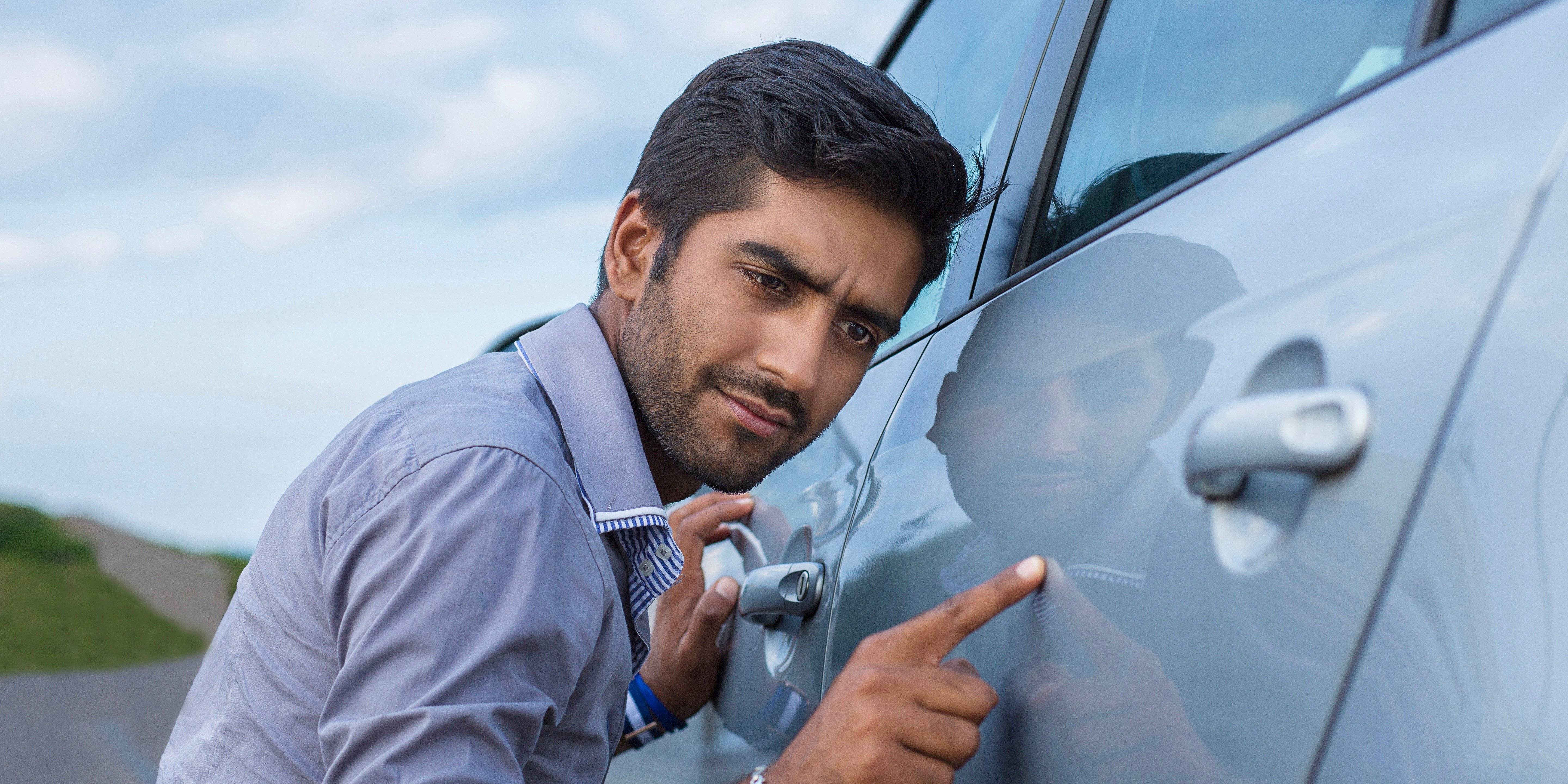 A man looks closely at the side of his vehicle while crouching down, looking concerned.