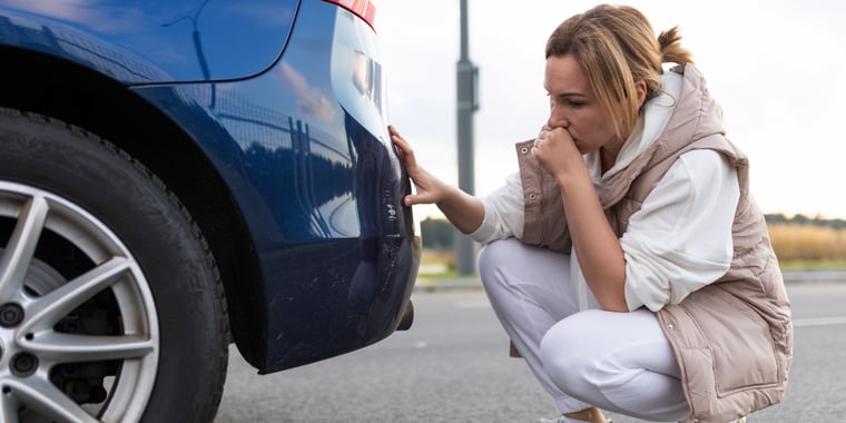 A woman crouches down next to the back of her car, looking for damage. The woman looks very worried.