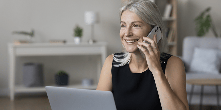 Picture of an older lady with her laptop open, smiling, and making a phone call on a smartphone.